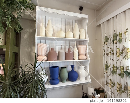 Pottery collection on white shelves in a cozy room surrounded by plants and curtains Pottery collection on white shelves in a cozy room surrounded by plants and curtains 141398291