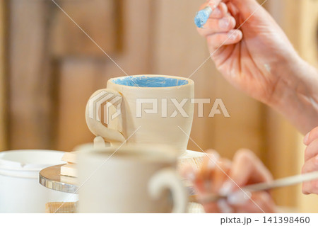 A woman steadily glazes a white ceramic mug on a rotating tray in a pottery studio; A woman steadily glazes a white ceramic mug on a rotating tray in a pottery studio; 141398460