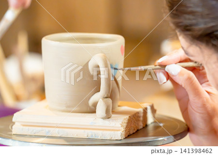 A woman steadily glazes a white ceramic mug on a rotating tray in a pottery studio; A woman steadily glazes a white ceramic mug on a rotating tray in a pottery studio; 141398462