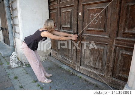 Woman leans against large aged wooden doors trying to open historic entrance with both hands. Heritage architecture, exploration, effort, tradition, travel, authentic atmosphere, lifestyle Woman leans against large aged wooden doors trying to open historic entrance with both hands. Heritage architecture, exploration, effort, tradition, travel, authentic atmosphere, lifestyle 141398891