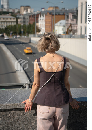 Woman observes Moscow city street from elevated pedestrian crossing in warm sunlight. Urban exploration, travel, transportation, modern lifestyle, city perspective, tourism 141398917