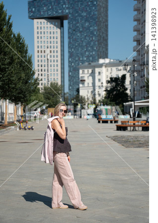 Side view of woman walking across sunny Shkolnaya Street in Moscow carrying jacket. Urban travel, lifestyle, warm weather, pedestrian zone, confidence, summer, freedom 141398923