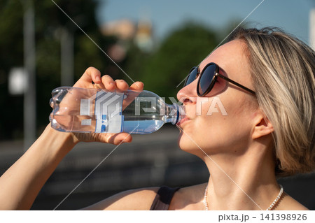 Woman drinking bottled water outdoors in Moscow. Hydration, summer heat, healthy lifestyle, wellbeing, refreshment, self care, fitness, urban living Woman drinking bottled water outdoors in Moscow. Hydration, summer heat, healthy lifestyle, wellbeing, refreshment, self care, fitness, urban living 141398926