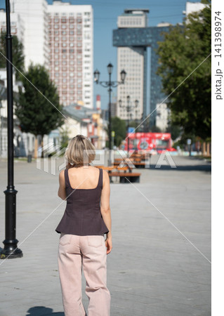 Back view of woman strolling along wide pedestrian Shkolnaya Street in Moscow during sunny day. Urban lifestyle, travel, freedom, architecture, city walk, modern living, wellbeing 141398974