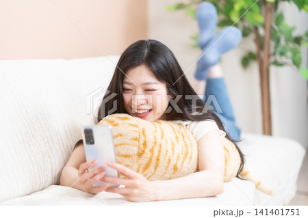 ソファーに寝転んでスマホを見る笑顔の若い女性 ソファーに寝転んでスマホを見る笑顔の若い女性 141401751