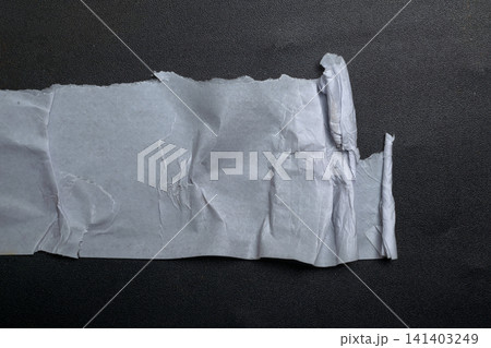 A piece of white, creased paper with jagged, torn edges resting on a dark, textured surface, creating a minimalist high-contrast composition for design use 141403249