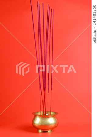 A close-up shot of several magenta incense sticks standing upright in a traditional polished brass bowl, set against a solid vibrant red background 141403250