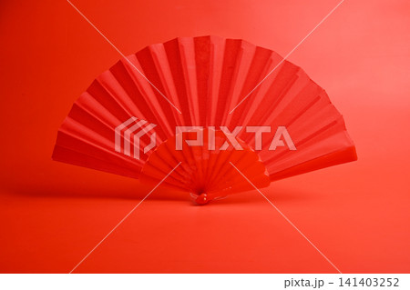 A close-up of a traditional red handheld folding fan isolated against a matching solid red background, representing elegance, culture, and minimal design A close-up of a traditional red handheld folding fan isolated against a matching solid red background, representing elegance, culture, and minimal design 141403252