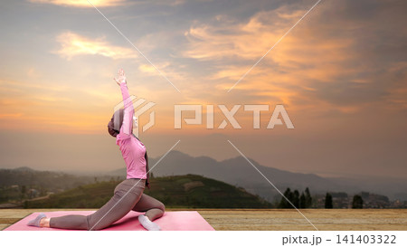 A young Indonesian southeast asian Muslim woman wearing a hijab practices yoga on a mat outdoors, stretching during a beautiful sunset with scenic mountain views A young Indonesian southeast asian Muslim woman wearing a hijab practices yoga on a mat outdoors, stretching during a beautiful sunset with scenic mountain views 141403322