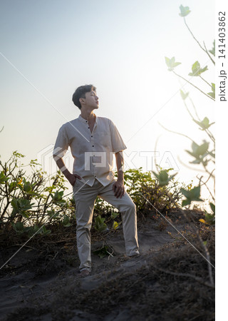 A handsome young Indonesian southeast asian man stands confidently on a sandy dune, wearing a casual linen shirt and trousers, backlit by the warm golden light of the sun A handsome young Indonesian southeast asian man stands confidently on a sandy dune, wearing a casual linen shirt and trousers, backlit by the warm golden light of the sun 141403862
