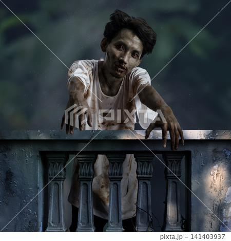 A terrifying zombie man with bloody makeup and torn clothes leaning over an old balcony railing, reaching his hands out in a dark horror scene for Horror or Halloween. Concept of Halloween A terrifying zombie man with bloody makeup and torn clothes leaning over an old balcony railing, reaching his hands out in a dark horror scene for Horror or Halloween. Concept of Halloween 141403937