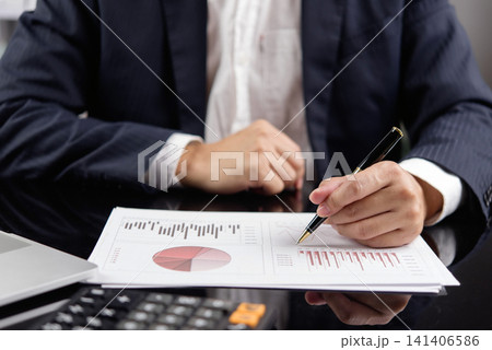 Businessman in navy suit holding financial report with bar chart and pie chart, using pen for data analysis. Business finance, investment, revenue, corporate strategy and financial planning concept. Businessman in navy suit holding financial report with bar chart and pie chart, using pen for data analysis. Business finance, investment, revenue, corporate strategy and financial planning concept. 141406586