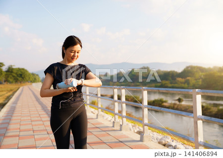 Young Asian woman checking her smartwatch while exercising at a riverside park Young Asian woman checking her smartwatch while exercising at a riverside park 141406894