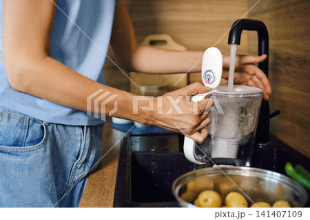 Unrecognizable woman draws water from tap into drinking water purification filter in kitchen Unrecognizable woman draws water from tap into drinking water purification filter in kitchen 141407109