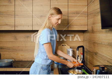 Woman stands at kitchen sink, washing carrots in kitchen Woman stands at kitchen sink, washing carrots in kitchen 141407111