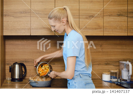 Young woman pours boiled vegetables into pot with parchment paper while cooking in kitchen 141407137