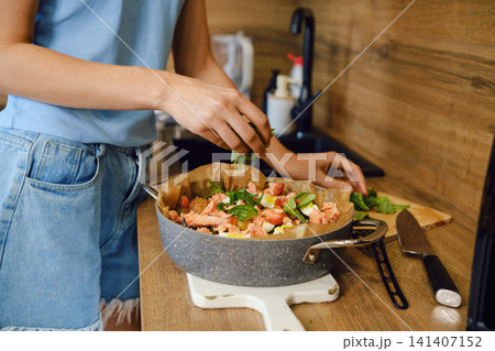 Woman adds basil to a pot with roasted vegetables and pink salmon Woman adds basil to a pot with roasted vegetables and pink salmon 141407152