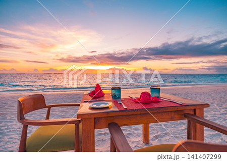 Dining table set for two on a beach terrace at sunset with the ocean beyond. Tropical resort restaurant at golden hour. Romantic honeymoon dinner travel background. Dining table set for two on a beach terrace at sunset with the ocean beyond. Tropical resort restaurant at golden hour. Romantic honeymoon dinner travel background. 141407299