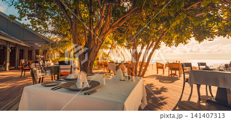 Romantic beach restaurant dining tables under trees at sunset warm lantern light tropical resort terrace. Perfect evening travel hospitality dining outdoors. Luxury honeymoon getaway ambience scenic Romantic beach restaurant dining tables under trees at sunset warm lantern light tropical resort terrace. Perfect evening travel hospitality dining outdoors. Luxury honeymoon getaway ambience scenic 141407313