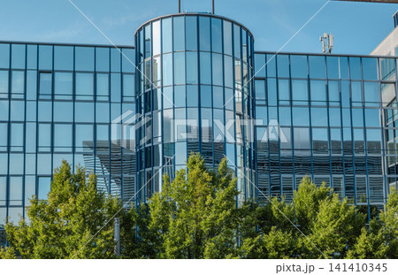 Modern glass office building facade near Hackerbruecke in Munich, Bavaria, with metal sunshade structure and green trees in foreground 141410345