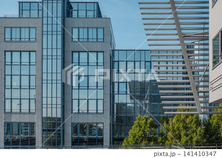 Modern glass office building facade near Hackerbruecke in Munich, Bavaria, with metal sunshade structure and green trees in foreground 141410347