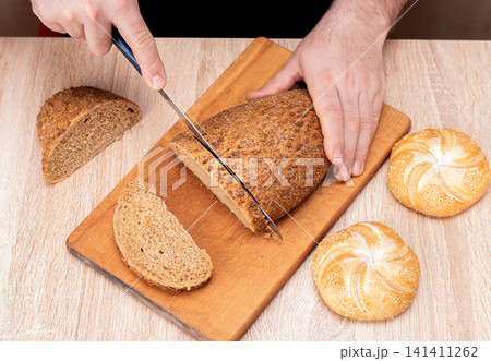 A man with a knife cuts bread on a wooden board. French baguettes. Different breeds on wooden background. A man with a knife cuts bread on a wooden board. French baguettes. Different breeds on wooden background. 141411262