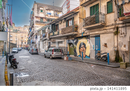 Naples, italy. December 30, 2024. Cobblestone street in naples, italy, featuring a large street art mural of footballer diego maradona on an old building wall Naples, italy. December 30, 2024. Cobblestone street in naples, italy, featuring a large street art mural of footballer diego maradona on an old building wall 141411895