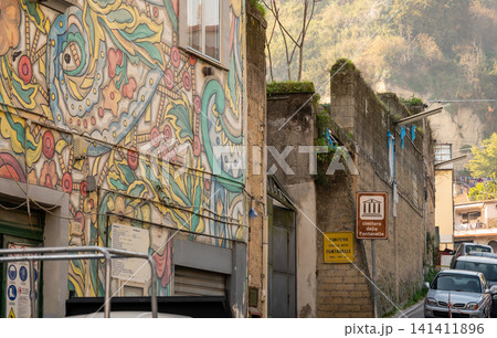 Naples, italy. December 30, 2024. Naples street scene featuring vibrant mural on building facade, alongside historic architecture and sign pointing to fontanelle cemetery Naples, italy. December 30, 2024. Naples street scene featuring vibrant mural on building facade, alongside historic architecture and sign pointing to fontanelle cemetery 141411896