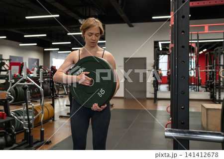Female athlete loading heavy weight plate onto barbell in squat rack inside bright modern gym, focused expression, strong posture, and visible effort during strength training 141412678