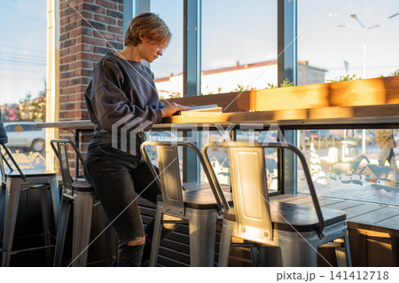 Young woman at cafe counter stirring hot drink beside window with phone, potted plants, casual cutout shoulder outfit, relaxed urban lifestyle mood in bright daylight Young woman at cafe counter stirring hot drink beside window with phone, potted plants, casual cutout shoulder outfit, relaxed urban lifestyle mood in bright daylight 141412718