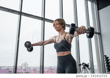 Athletic woman lifting dumbbells in lateral raise exercise inside bright modern gym with floor to ceiling windows and city skyline, focused posture and strong toned arms Athletic woman lifting dumbbells in lateral raise exercise inside bright modern gym with floor to ceiling windows and city skyline, focused posture and strong toned arms 141412725