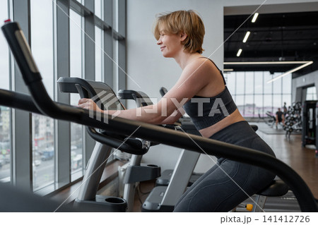 Focused woman exercising on treadmill in modern gym with floor to ceiling windows, city skyline outside, leaning forward on console in side profile, bright natural light and calm concentration Focused woman exercising on treadmill in modern gym with floor to ceiling windows, city skyline outside, leaning forward on console in side profile, bright natural light and calm concentration 141412726