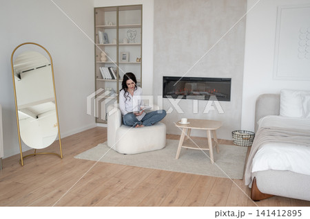 Relaxing at home, woman reading magazine on cozy white chair, drinking morning coffee, enjoying leisure time. Lifestyle, relaxation, comfort, tranquility, indoors Relaxing at home, woman reading magazine on cozy white chair, drinking morning coffee, enjoying leisure time. Lifestyle, relaxation, comfort, tranquility, indoors 141412895