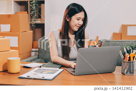 Startup SME small business entrepreneur of freelance Asian woman wearing apron using laptop and box to receive and review orders online to prepare to pack sell to customers, online sme business ideas. Startup SME small business entrepreneur of freelance Asian woman wearing apron using laptop and box to receive and review orders online to prepare to pack sell to customers, online sme business ideas. 141412902
