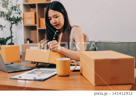 Startup SME small business entrepreneur of freelance Asian woman wearing apron using laptop and box to receive and review orders online to prepare to pack sell to customers, online sme business ideas. Startup SME small business entrepreneur of freelance Asian woman wearing apron using laptop and box to receive and review orders online to prepare to pack sell to customers, online sme business ideas. 141412912