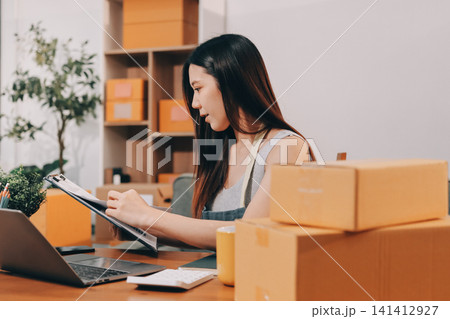Startup SME small business entrepreneur of freelance Asian woman wearing apron using laptop and box to receive and review orders online to prepare to pack sell to customers, online sme business ideas. Startup SME small business entrepreneur of freelance Asian woman wearing apron using laptop and box to receive and review orders online to prepare to pack sell to customers, online sme business ideas. 141412927