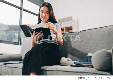 Freelance Asian woman holding mobile phone typing on laptop and working from home. Happy young girl sitting on sofa in living room. 141412979
