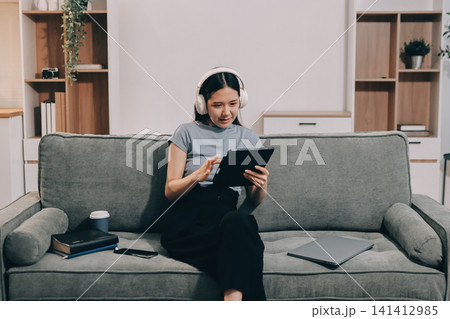 Freelance Asian woman holding mobile phone typing on laptop and working from home. Happy young girl sitting on sofa in living room. 141412985