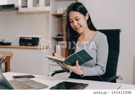 Young asian businesswoman is smiling and working on a laptop while sitting at her desk in a modern office Young asian businesswoman is smiling and working on a laptop while sitting at her desk in a modern office 141412996