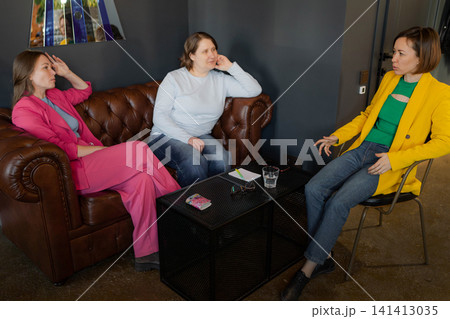 Two women holding a conversation in chic cafe lounge on brown leather sofa with books in the background. Cafe, lounge, conversation, leather sofa, books. 141413035