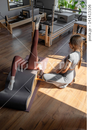 Woman doing exercises on back spine corrector with trainer. Reformer pilates studio machine for fitness workouts in gym. Fit, healthy and strong authentical body. Fitness concept Woman doing exercises on back spine corrector with trainer. Reformer pilates studio machine for fitness workouts in gym. Fit, healthy and strong authentical body. Fitness concept 141413106