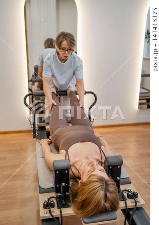 Female instructor guiding client through leg extension routine on pilates reformer in airy gym. Leg extension, pilates reformer, instructor, routine, gym Female instructor guiding client through leg extension routine on pilates reformer in airy gym. Leg extension, pilates reformer, instructor, routine, gym 141413175