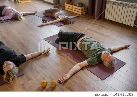 Senior gray-haired adult man doing pilates exercises for back and joint on a mat. A trainer helps him gently recover Senior gray-haired adult man doing pilates exercises for back and joint on a mat. A trainer helps him gently recover 141413202