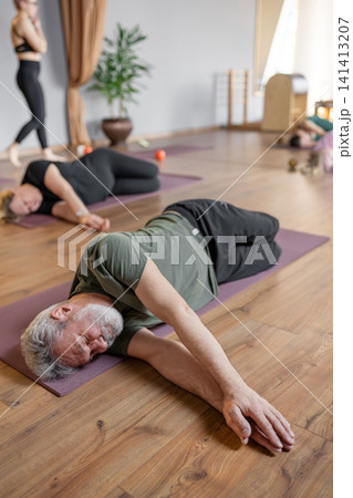 Senior gray-haired adult man doing pilates exercises for back and joint on a mat. A trainer helps him gently recover Senior gray-haired adult man doing pilates exercises for back and joint on a mat. A trainer helps him gently recover 141413207