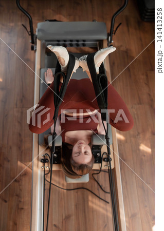Woman holding high leg stretch with instructor's assistance on pilates reformer in neutral studio. High leg stretch, pilates reformer, assistance, instructor, studio. Woman holding high leg stretch with instructor's assistance on pilates reformer in neutral studio. High leg stretch, pilates reformer, assistance, instructor, studio. 141413258
