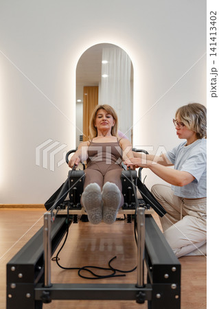 Sporty mature woman practicing pilates abs routine on reformer machine in daylight studio with trainer. Pilates abs, reformer machine, sporty, studio, routine Sporty mature woman practicing pilates abs routine on reformer machine in daylight studio with trainer. Pilates abs, reformer machine, sporty, studio, routine 141413402