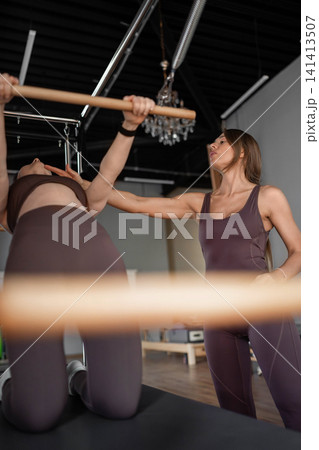 Elegant pilates pair performing synchronized deep stretches using reformer beds in bright wood-floored studio. Synchronized, deep stretch, reformer, studio, wood floor 141413507