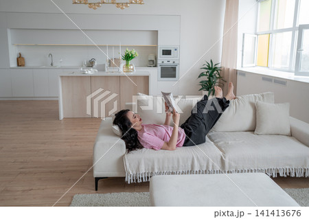 Woman relaxing at home with magazine on cozy white sofa, enjoying leisure time, minimalist kitchen in the background. Lifestyle, relaxation, comfort, tranquility, indoors 141413676