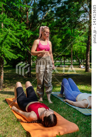 Two women in group yoga class resting on mats with instructor in summer park. Resting, yoga, mats, instructor, group, park, summer. 141413685