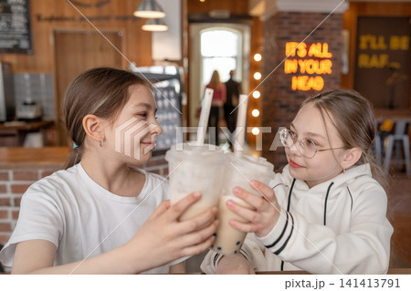 Girls sitting side by side at cafe table raising milkshake cups and smiling in warm light. Milkshake, cafe, girls, smiling, table. Girls sitting side by side at cafe table raising milkshake cups and smiling in warm light. Milkshake, cafe, girls, smiling, table. 141413791